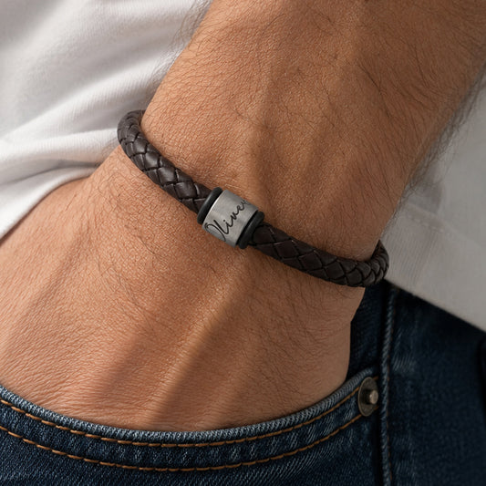 A close-up of a man's wrist wearing a dark brown braided leather bracelet with a silver ring engraved with a child's name. 
