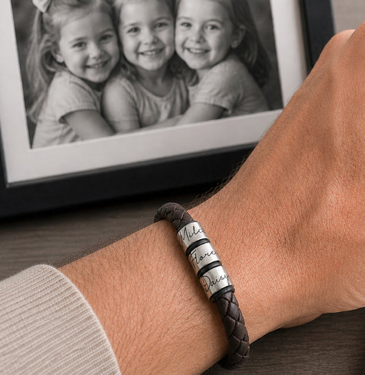 A close-up of a man's hand wearing a dark brown braided leather bracelet with three silver rings engraved with names, sitting at a desk next to a framed photo of children.