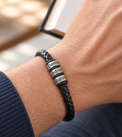 Close-up of a man's wrist wearing a personalised black braided leather bracelet with three silver-coloured rings engraved with names.